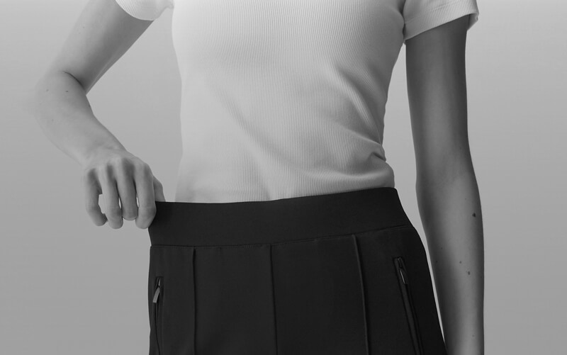 Gros plan en noir et blanc d'une personne portant un t-shirt blanc côtelé et un pantalon OPUS noir. Le cadre capture le torse de la taille à la poitrine, avec la main de la personne reposant sur sa taille. En raison du pont de taille, le pantalon s'ajuste très bien.
